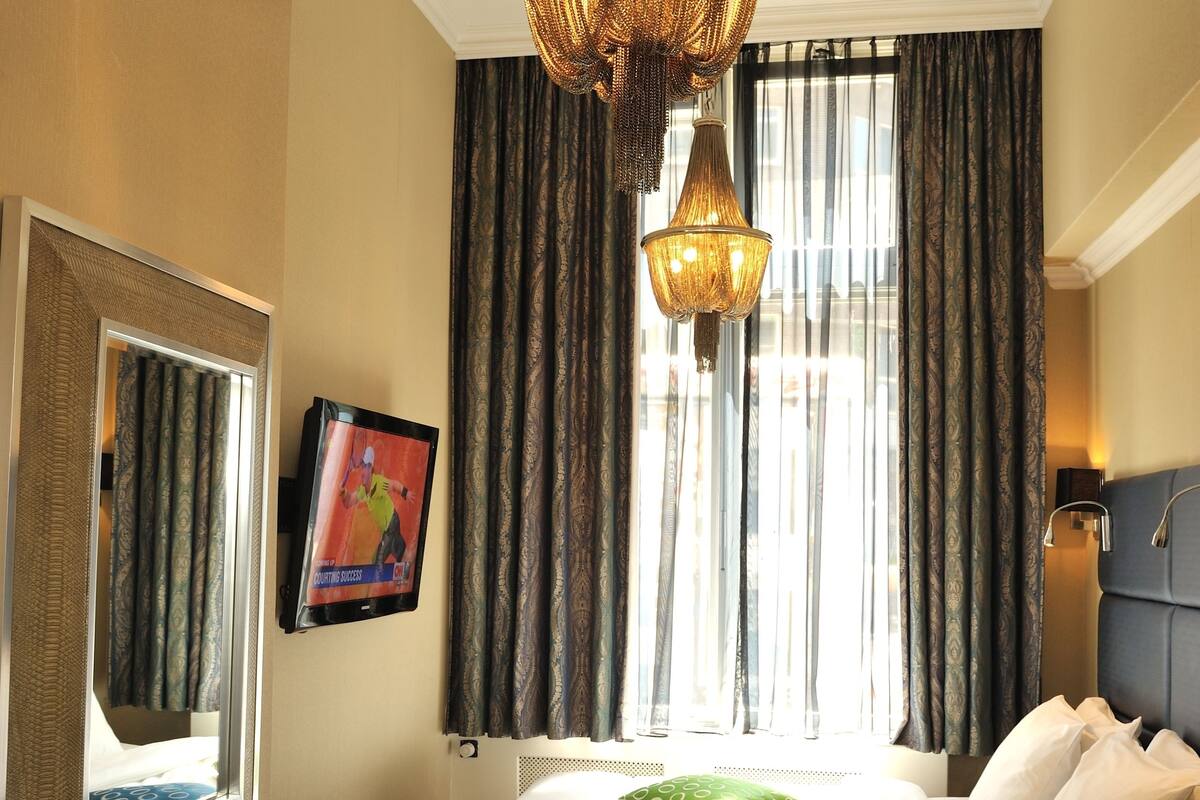 A hotel room with a bed, a desk with a plate of apples, a chair, a small table with a bottle, a hanging light fixture, a mirror, and a window with curtains.