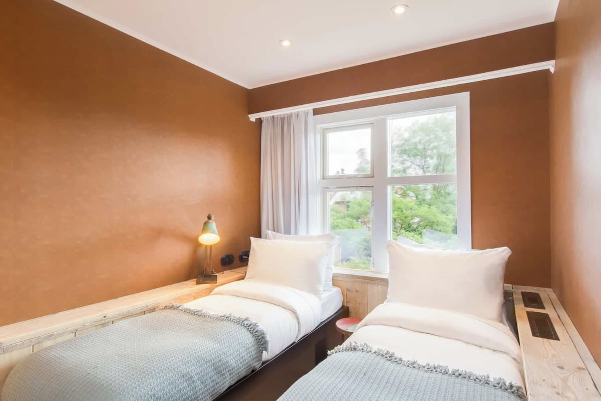 A room with two beds, a window with curtains, and a bedside lamp.