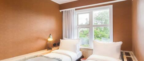 A room with two beds, a window with curtains, and a bedside lamp.