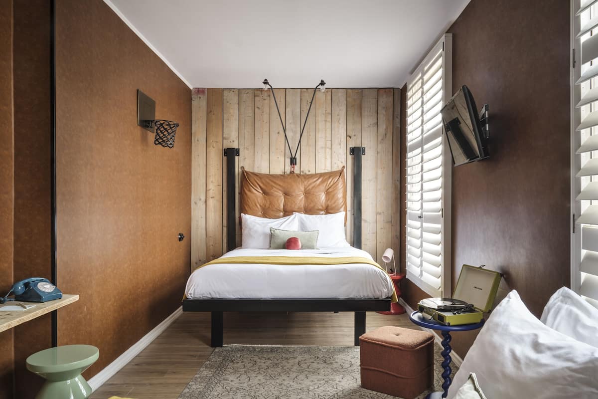 A bedroom with a bed, a bedside table, a lamp, a TV, and a window with blinds.