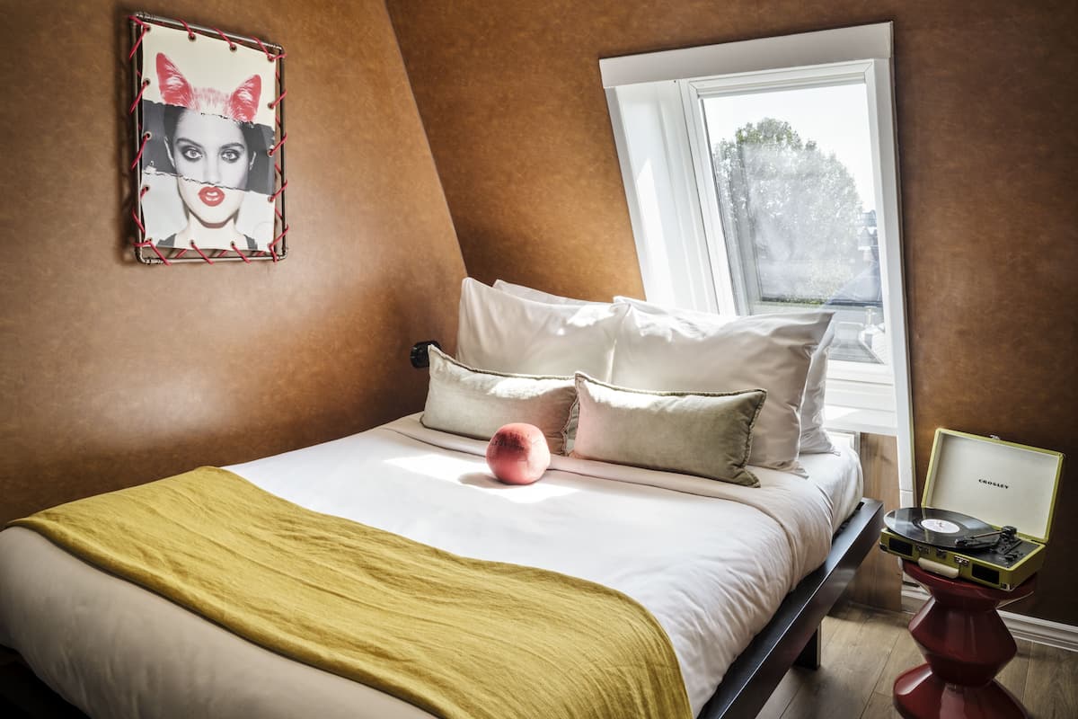 A bedroom with a bed, pillows, a bedside table with a record player, a window, and a wall-mounted picture.