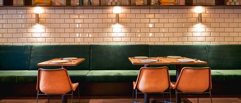 A restaurant with a green cushioned bench, two tables with chairs, and wall-mounted lights.