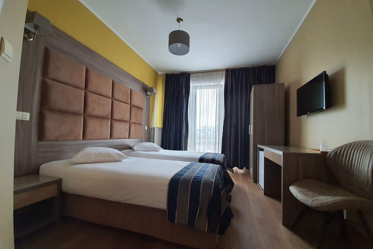Habitación de hotel con dos camas, un escritorio, una silla, un televisor y un ventanal con cortinas.