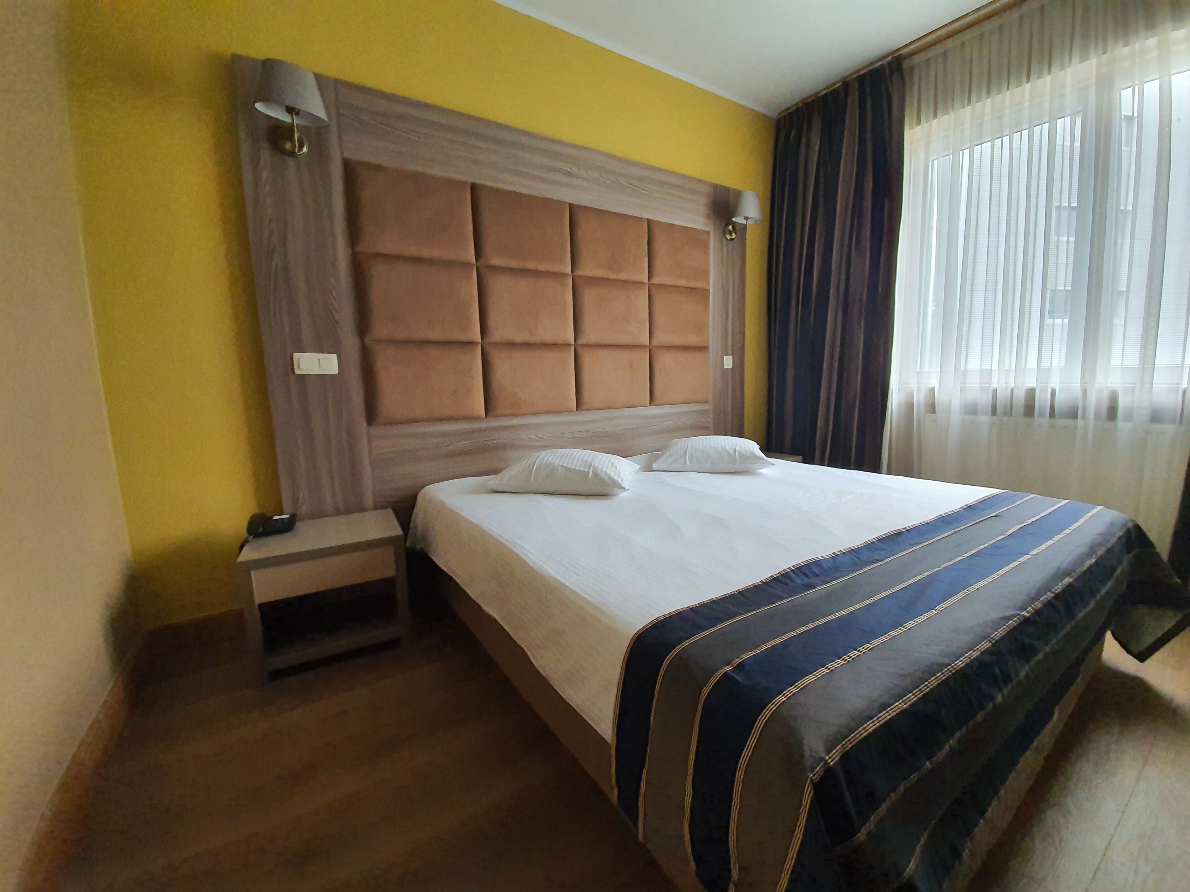 Una habitación de hotel con cama, mesita de noche, lámpara y ventana con cortinas.