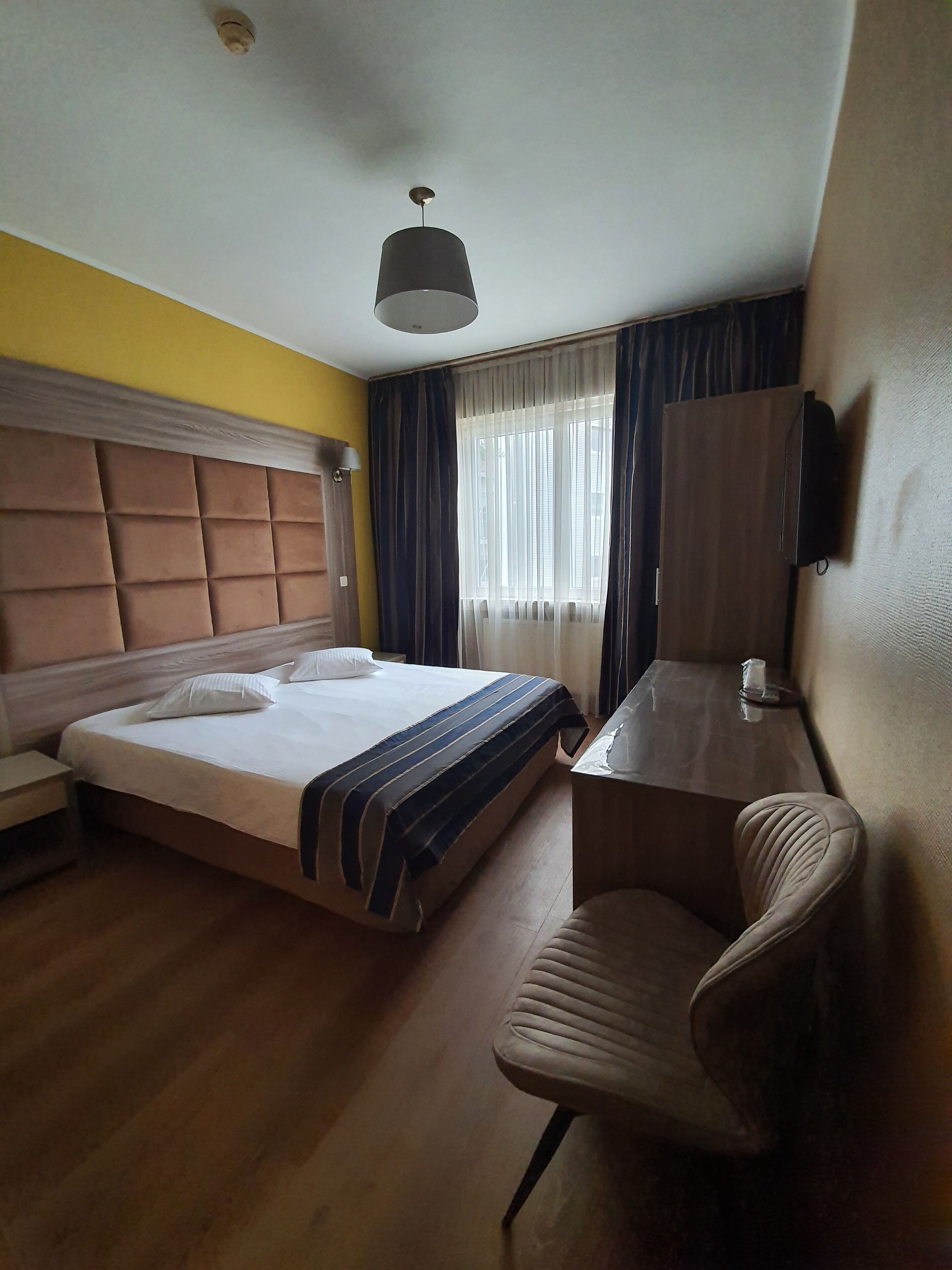 A hotel room with a bed, a desk, a chair, a wardrobe, and a ceiling light.