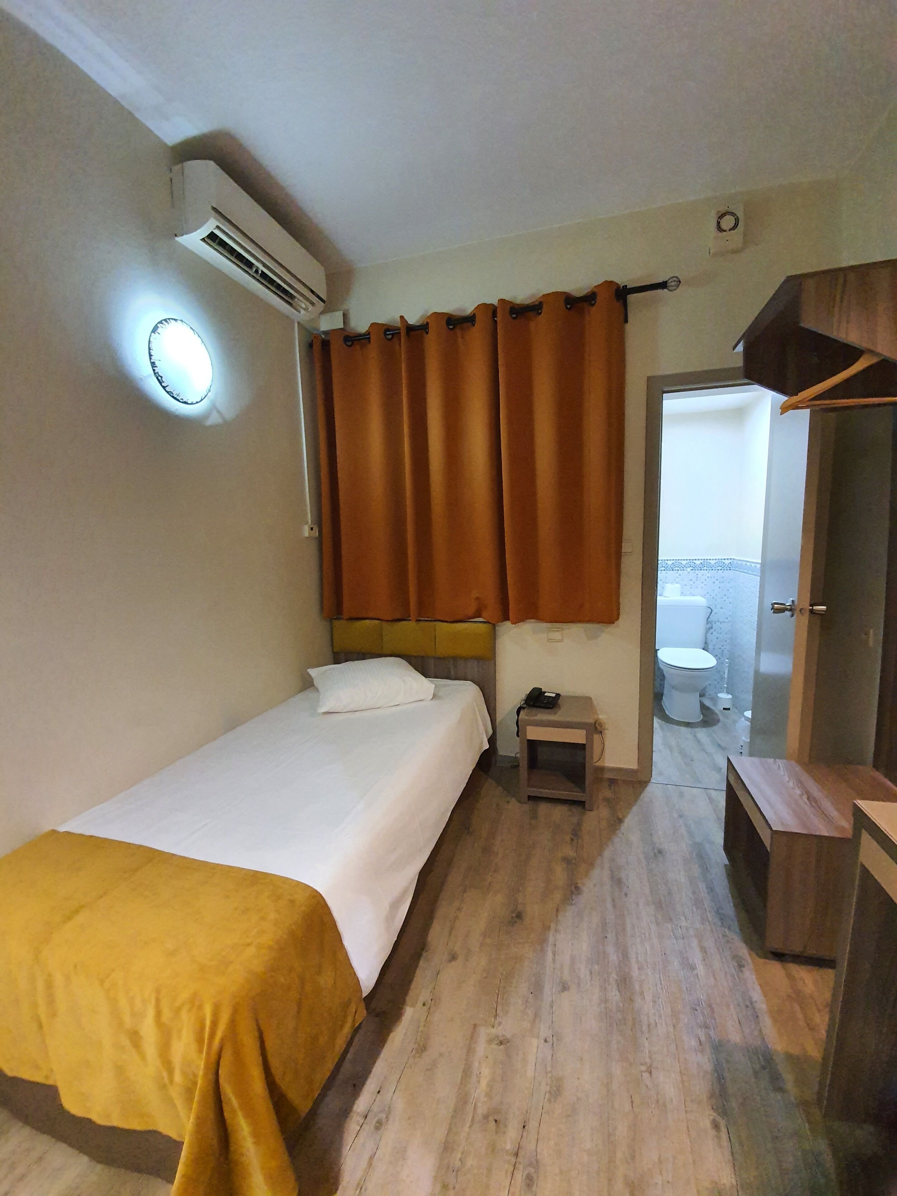 Una habitación de hotel con una cama, una mesita de noche con teléfono, un armario pequeño, un espejo y un baño visible a través de una puerta abierta.