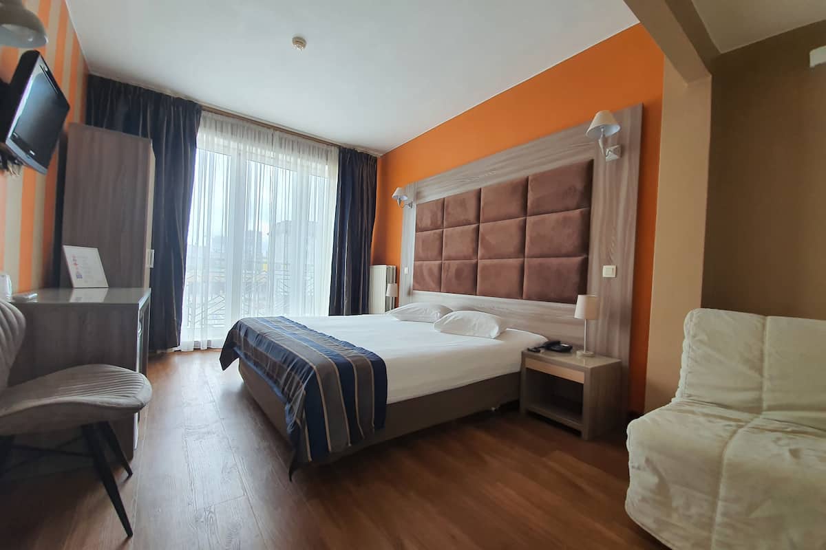 Habitación de hotel con una cama grande, un escritorio, una silla, un televisor montado en la pared y una ventana con cortinas.