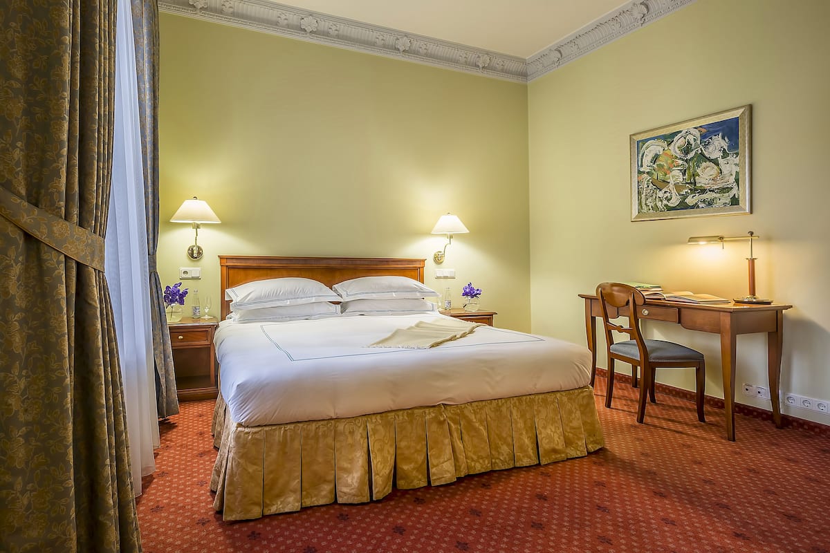 A hotel room with a large bed, two bedside lamps, a desk with a chair, and a painting on the wall.