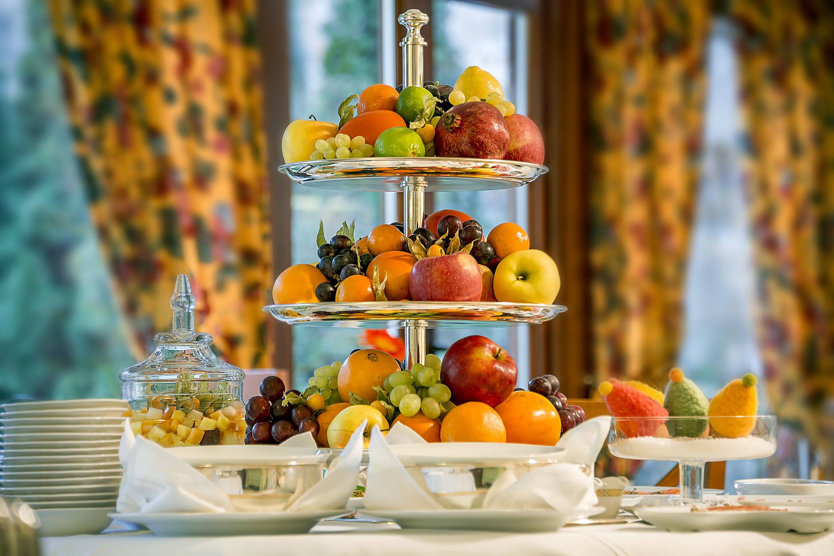 A three-tiered fruit stand with various fruits and a glass container of nuts.