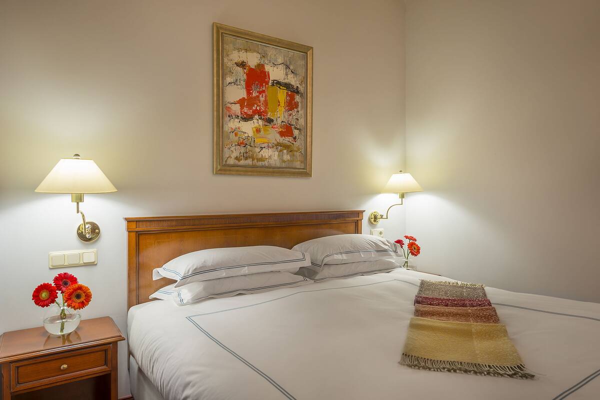 A hotel room with a wooden headboard, white bedding, two bedside lamps, a framed abstract painting, and a vase with flowers on the nightstand.