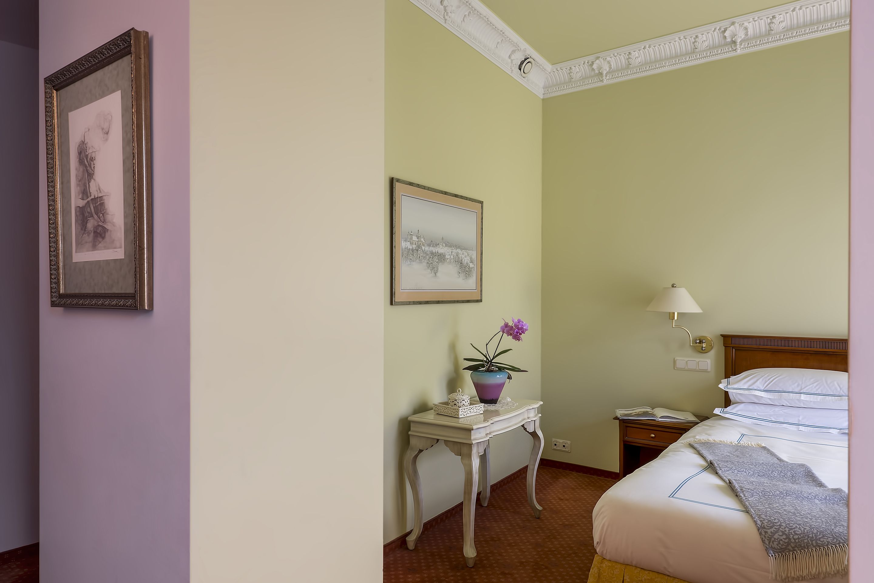 A hotel room with a bed, bedside table with a lamp, a small table with a vase of flowers, and two framed pictures on the wall.