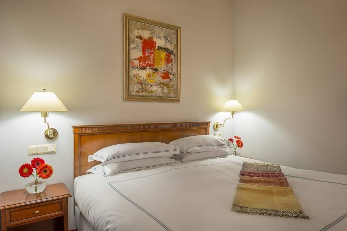 A hotel room with a wooden headboard, two bedside lamps, a painting on the wall, a bed with white linens, a nightstand with a vase of flowers, and a throw blanket on the bed.