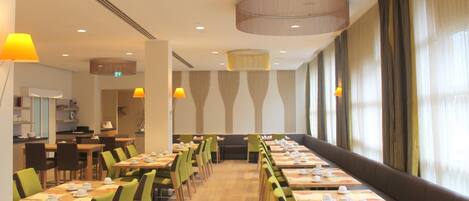 A restaurant with long tables and chairs, a large window with curtains, and a ceiling with recessed lighting.