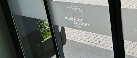A sign for "Cityhotel Kurfürst Balduin Garni" with a view of a paved street and greenery outside.