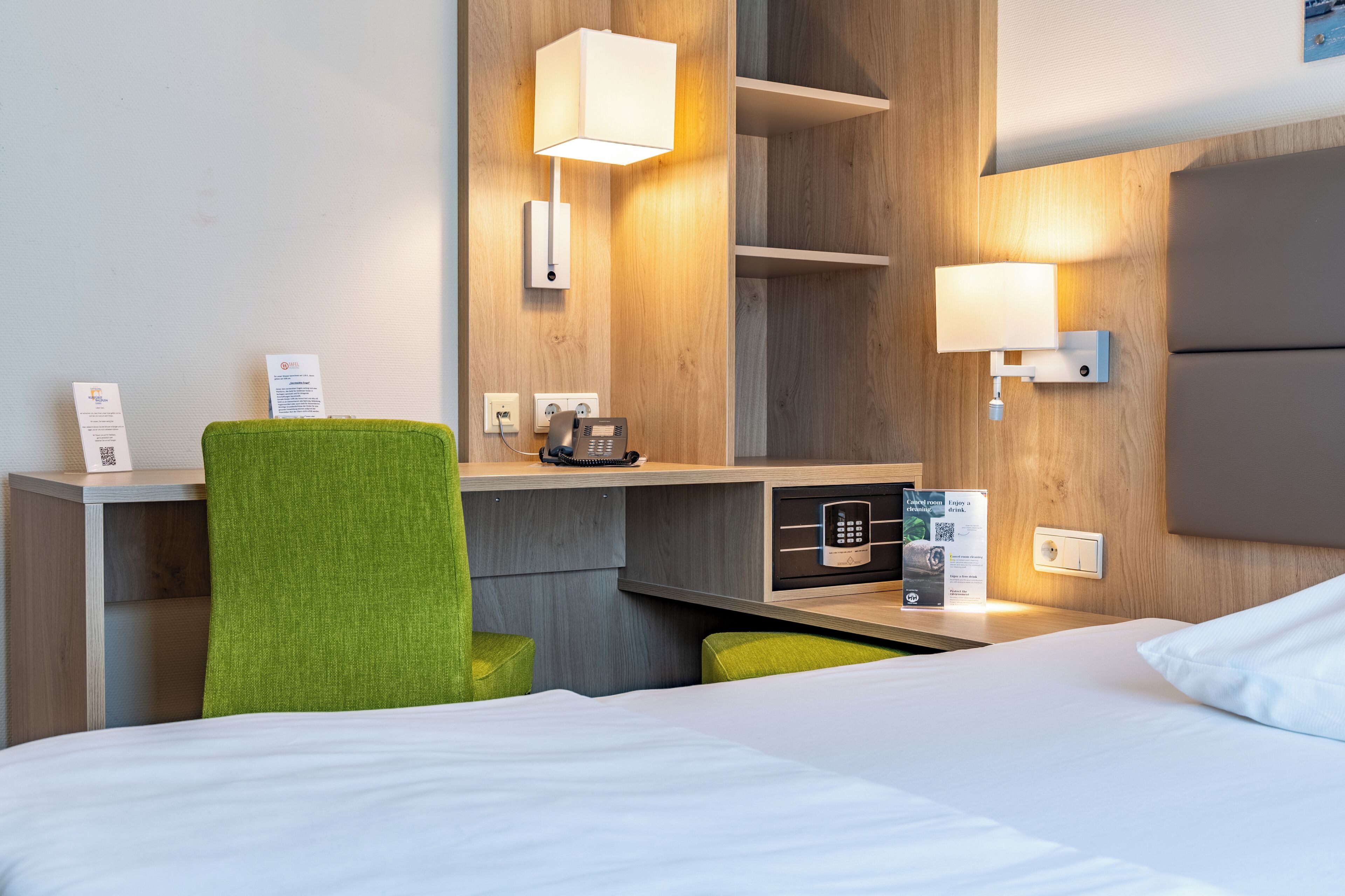 A hotel room with a bed, a desk with a telephone, a lamp, a mirror, and a green chair.