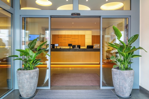 Zona de recepción con puerta de cristal, mostrador de recepción y dos plantas grandes en macetas.