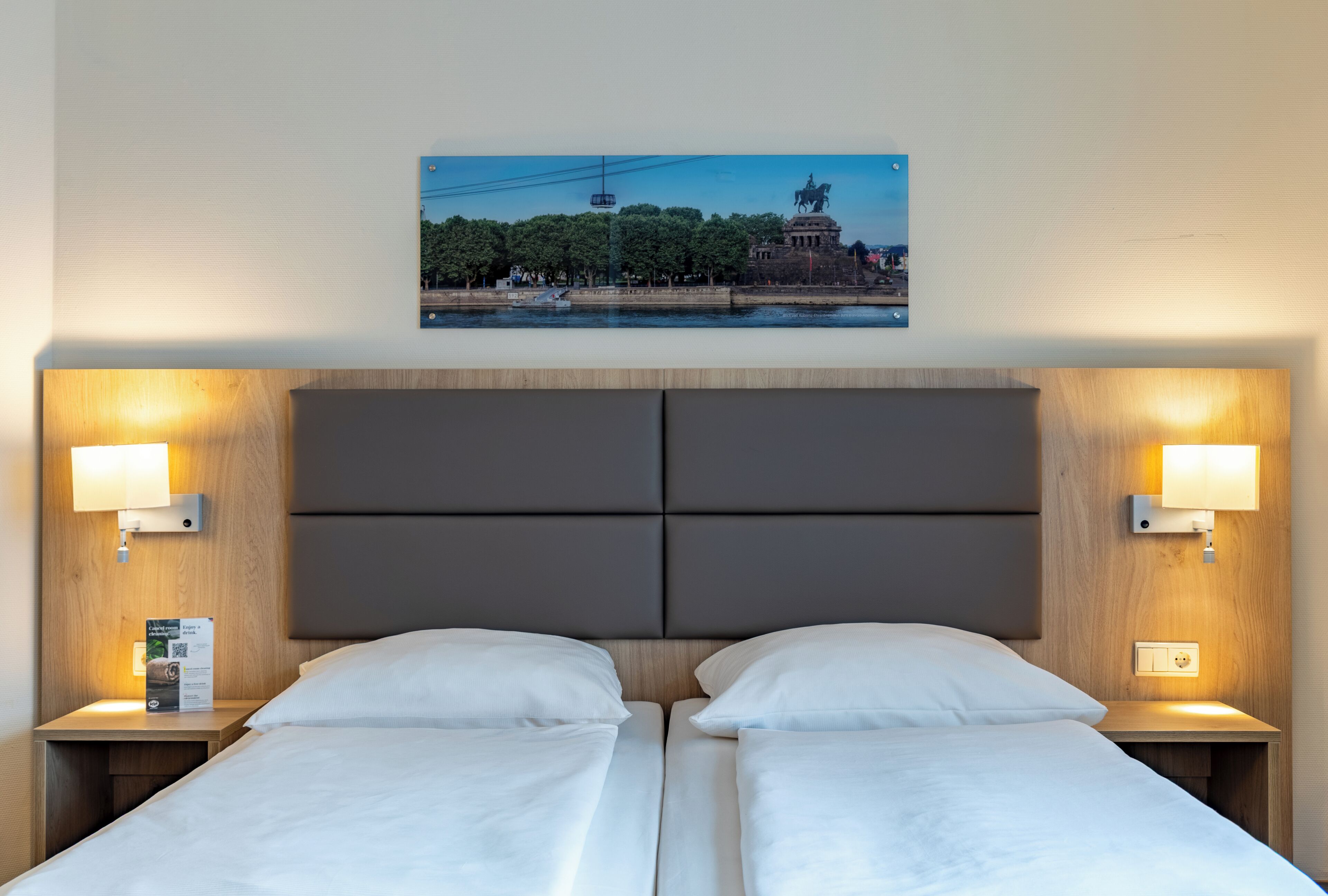 A hotel room with two beds, a large headboard, and a framed picture of a statue and building on the wall.