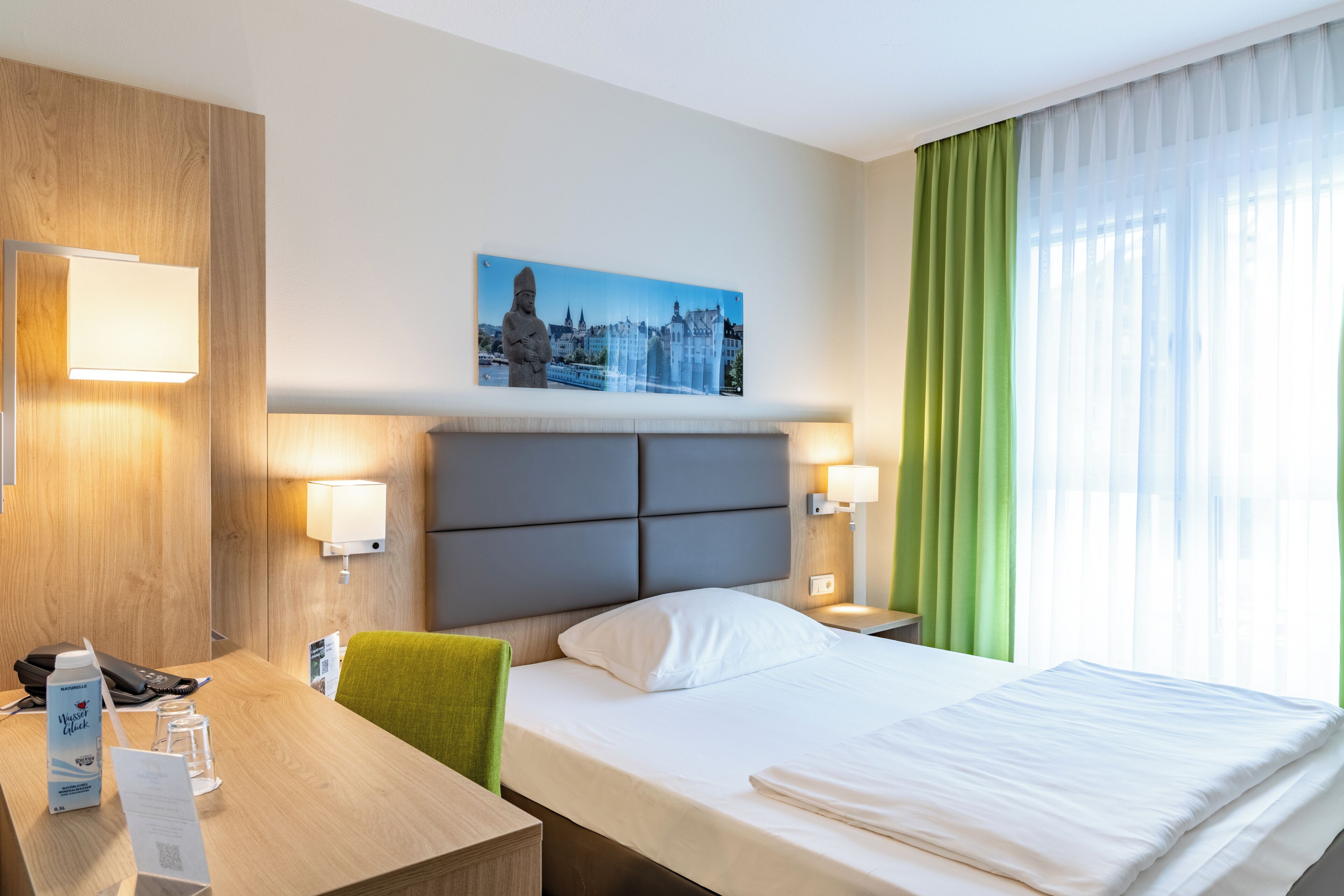 A hotel room with a bed, a desk with a telephone and a water bottle, a painting of a cityscape, and a window with green curtains.