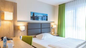 A hotel room with a bed, a desk with a telephone and a water bottle, a painting of a cityscape, and a window with green curtains.