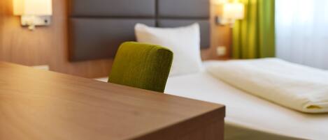 A hotel room with a wooden desk, a green chair, a bed with white bedding, and two wall-mounted lamps.