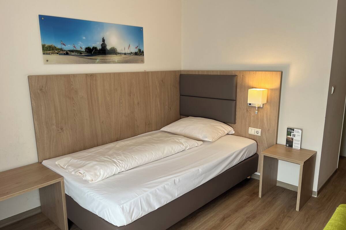 Un lit simple avec une tête de lit en bois, une table de chevet avec une lampe et un tableau encadré au mur.