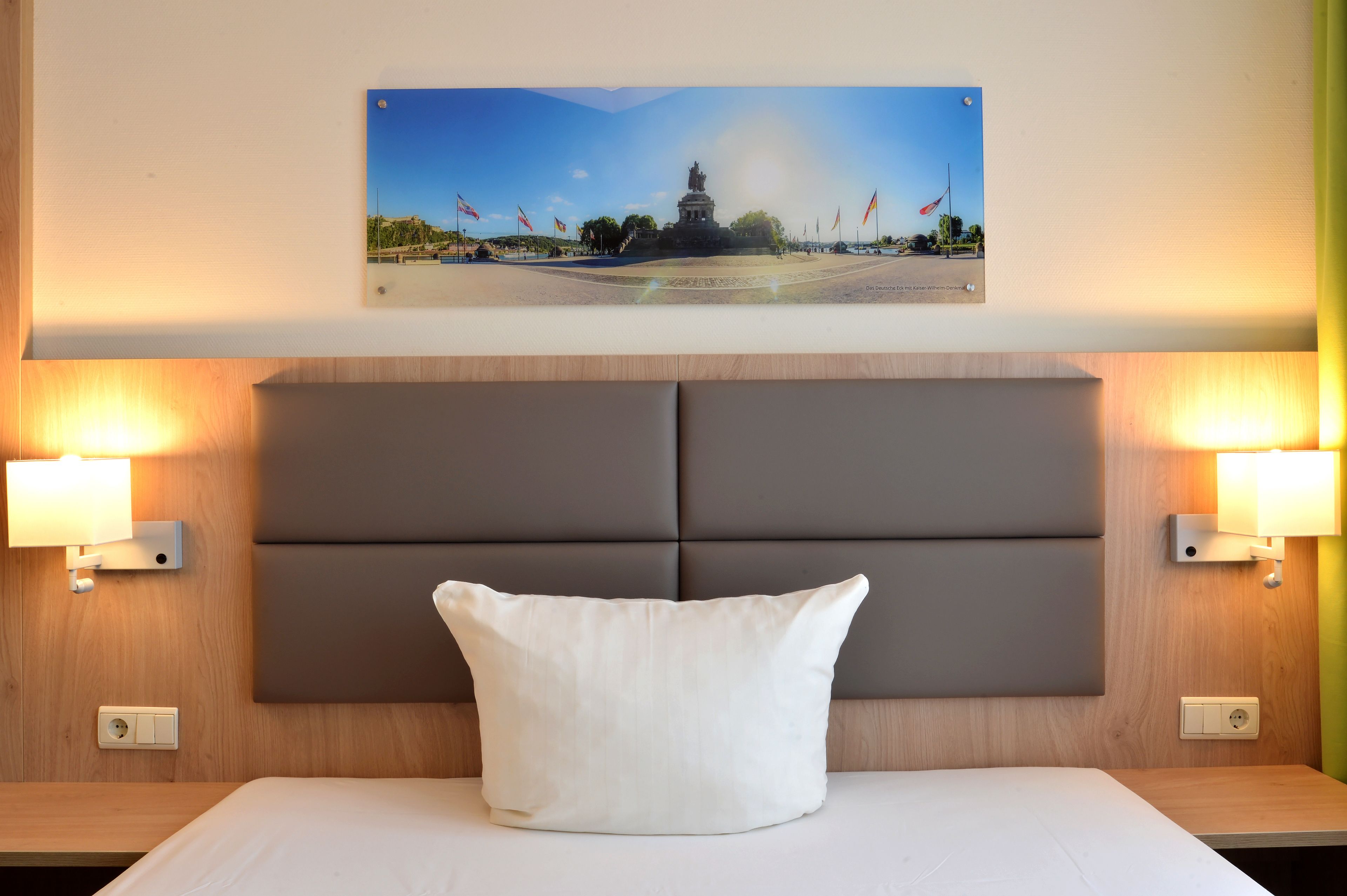 A hotel room with a bed, a headboard, two wall-mounted lamps, and a framed picture on the wall.