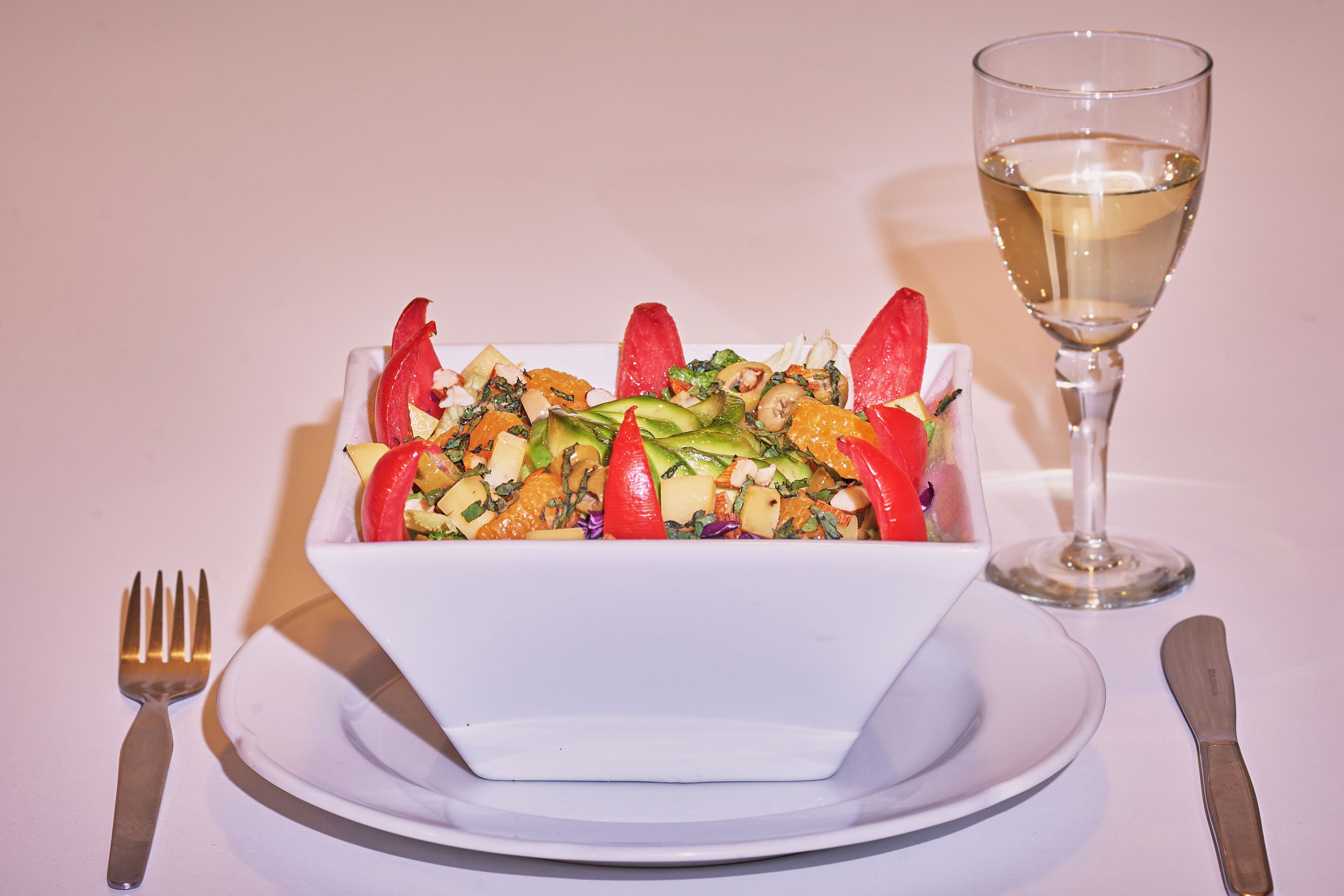 Uma salada em uma tigela branca com um copo de vinho branco sobre uma mesa.