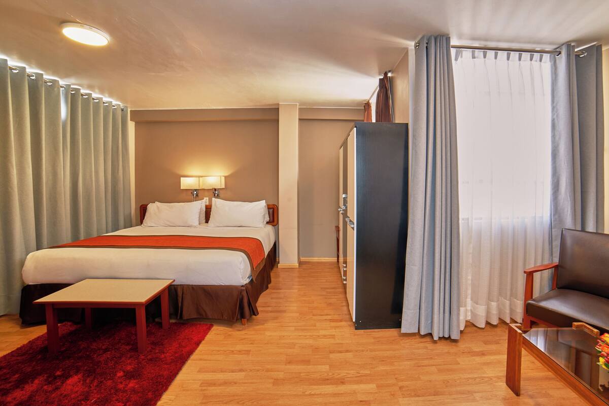 Ein Hotelzimmer mit Bett, Schreibtisch, Stuhl, Kommode und einem Fenster mit Vorhängen.