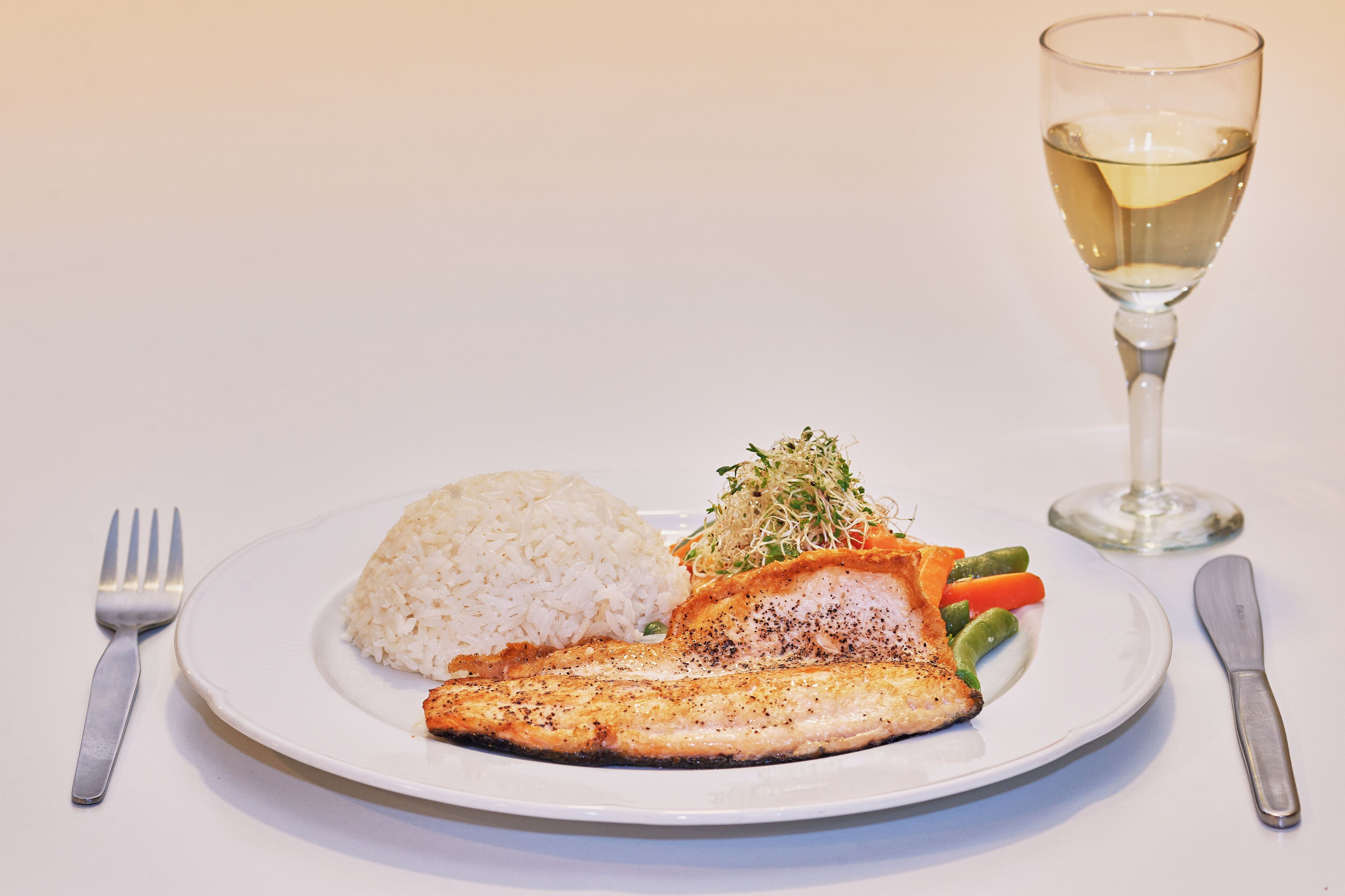 Une assiette de riz blanc, un morceau de poisson grillé et des légumes, accompagnés d’un verre de vin blanc et de couverts sur une table blanche.