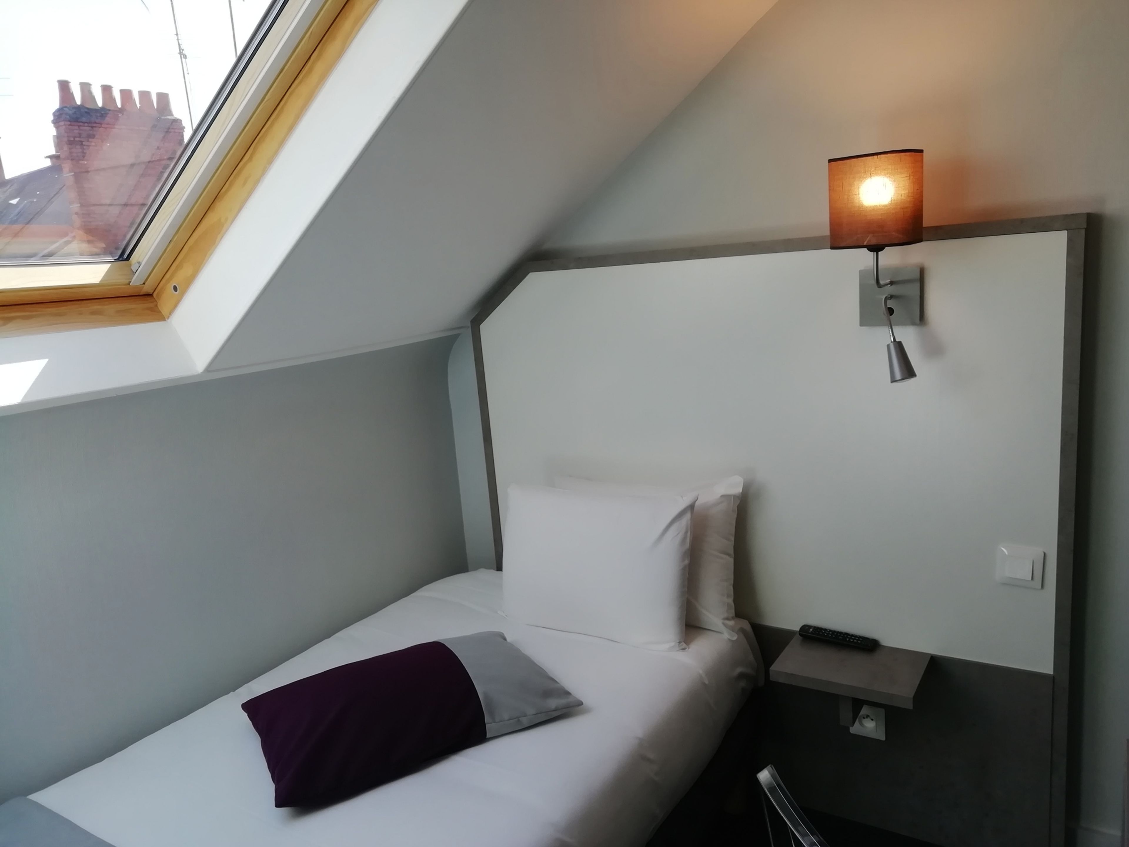 A small, modern hotel room with a single bed, a nightstand, a lamp, and a view of the outside through a skylight.
