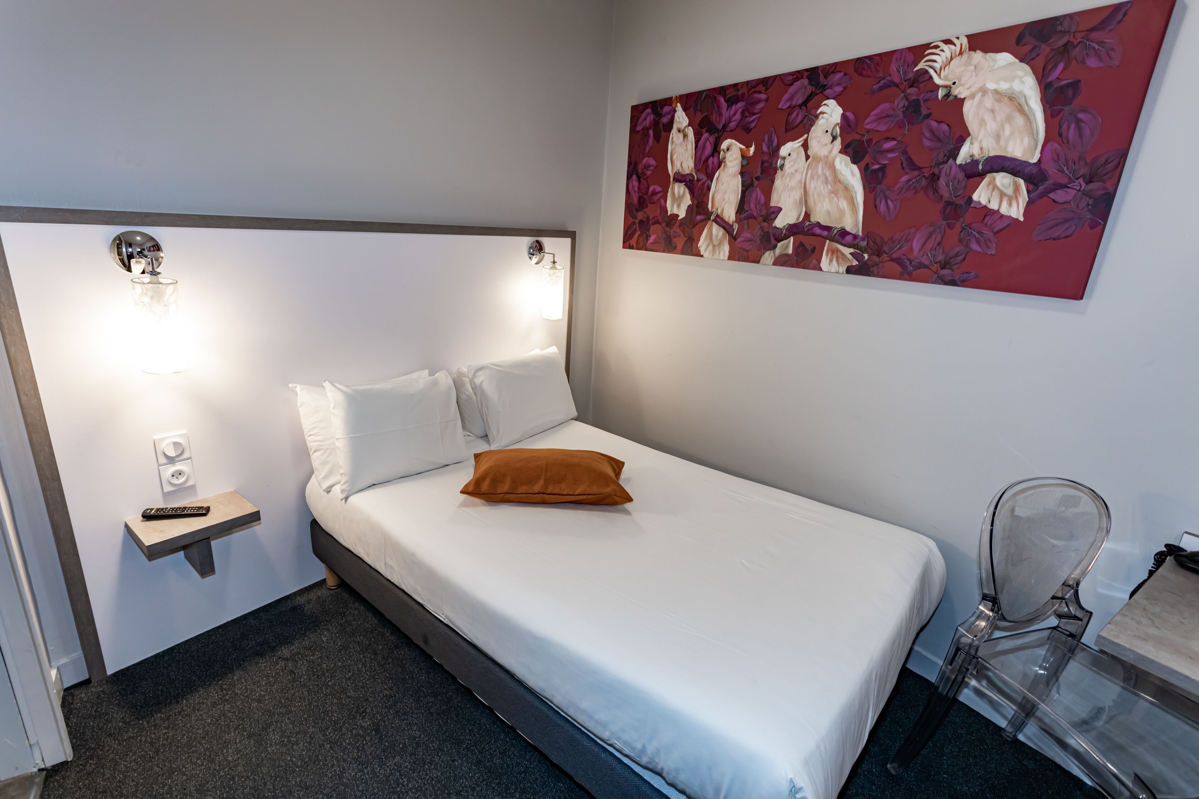 Una cama bien hecha con sábanas blancas y una almohada marrón, una mesita de noche con un control remoto, una lámpara de pared y un cuadro de pájaros en la pared.