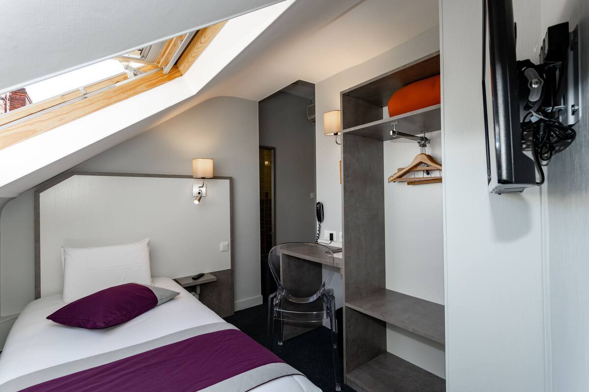 Une chambre d’hôtel avec une lucarne, un lit avec un oreiller violet, un bureau avec une chaise et une armoire avec un sac suspendu.
