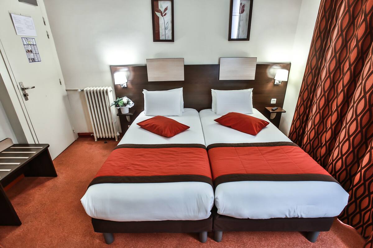 Une chambre d’hôtel avec deux lits, chacun doté d’une literie rouge et blanche, d’une tête de lit en bois, de lampes de chevet, d’un radiateur et de rideaux à motifs.