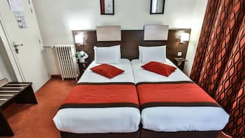 A hotel room with two beds, each with red and white bedding, a wooden headboard, bedside lamps, a radiator, and a patterned curtain.