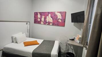 A hotel room with a bed, a desk with a chair, a flat-screen TV, a painting on the wall, and a ceiling light.