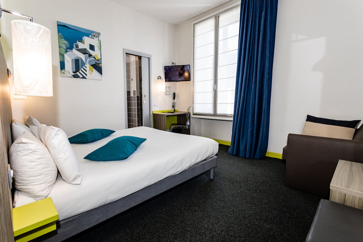 Une chambre d’hôtel avec un lit, un bureau, une chaise, une télévision et une fenêtre avec des rideaux.