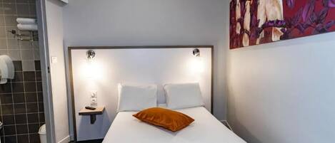 Una habitación de hotel con una cama, una mesita de noche, un cuadro en la pared y un baño visible a través de una puerta abierta.