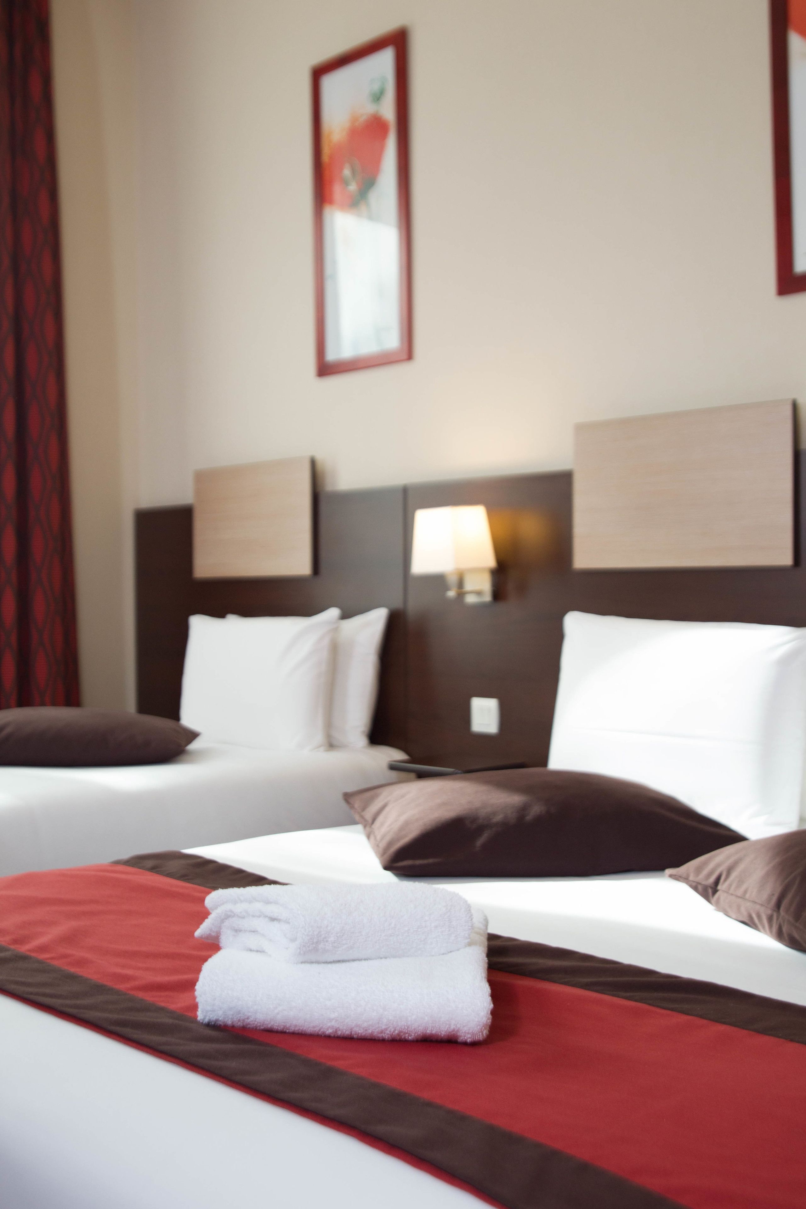 Une chambre d’hôtel avec deux lits, une couvre-lit rouge et marron, des oreillers blancs et un tableau encadré au mur.