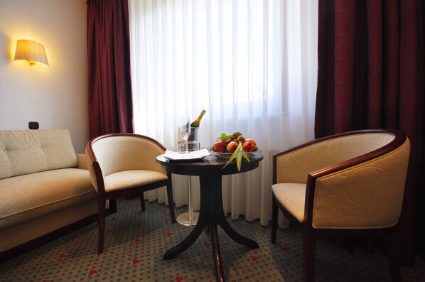 Habitación de hotel con sofá, dos sillones, una mesita con una botella de champán, una copa y frutas.