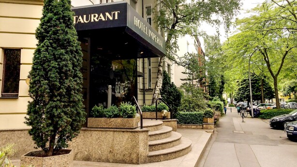 Un hôtel doté d’un restaurant au rez-de-chaussée, avec un escalier en pierre menant à l’entrée, le tout entouré de verdure et de voitures garées.