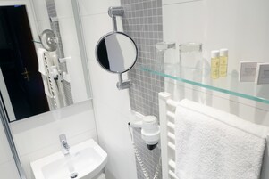 Un baño moderno con cabina de ducha de vidrio, un espejo redondo, un lavabo blanco y una estantería de vidrio con artículos de aseo.