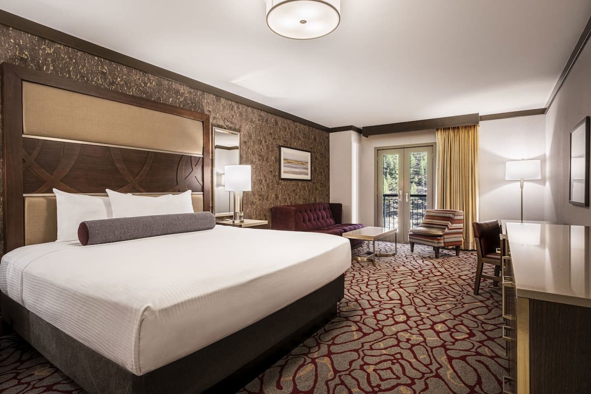 A hotel room with a large bed, a desk, a chair, a sofa, and a balcony with curtains.