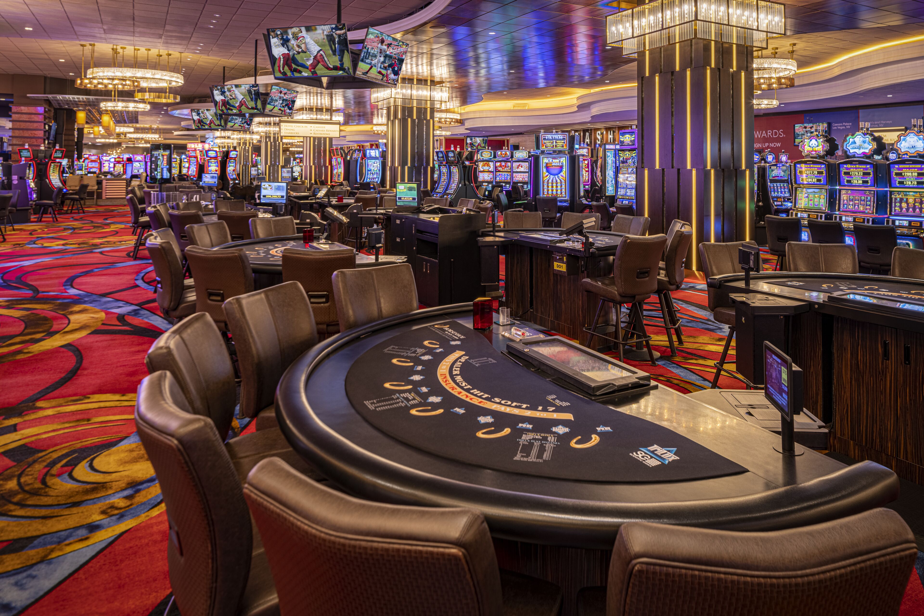 Un salón de casino con varias mesas de juego, máquinas tragamonedas y una pantalla grande mostrando un evento deportivo.