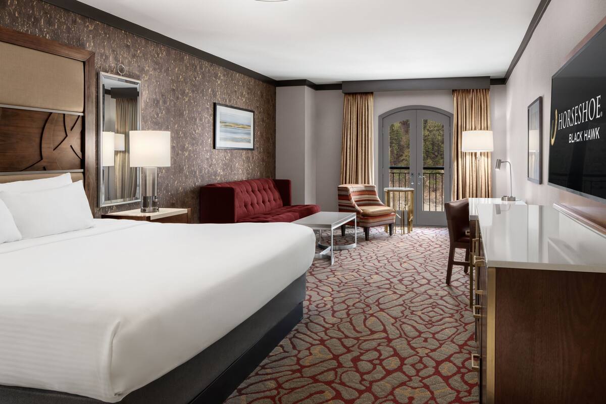 A hotel room with a large bed, a red sofa, a small table, a chair, a desk, and a TV.