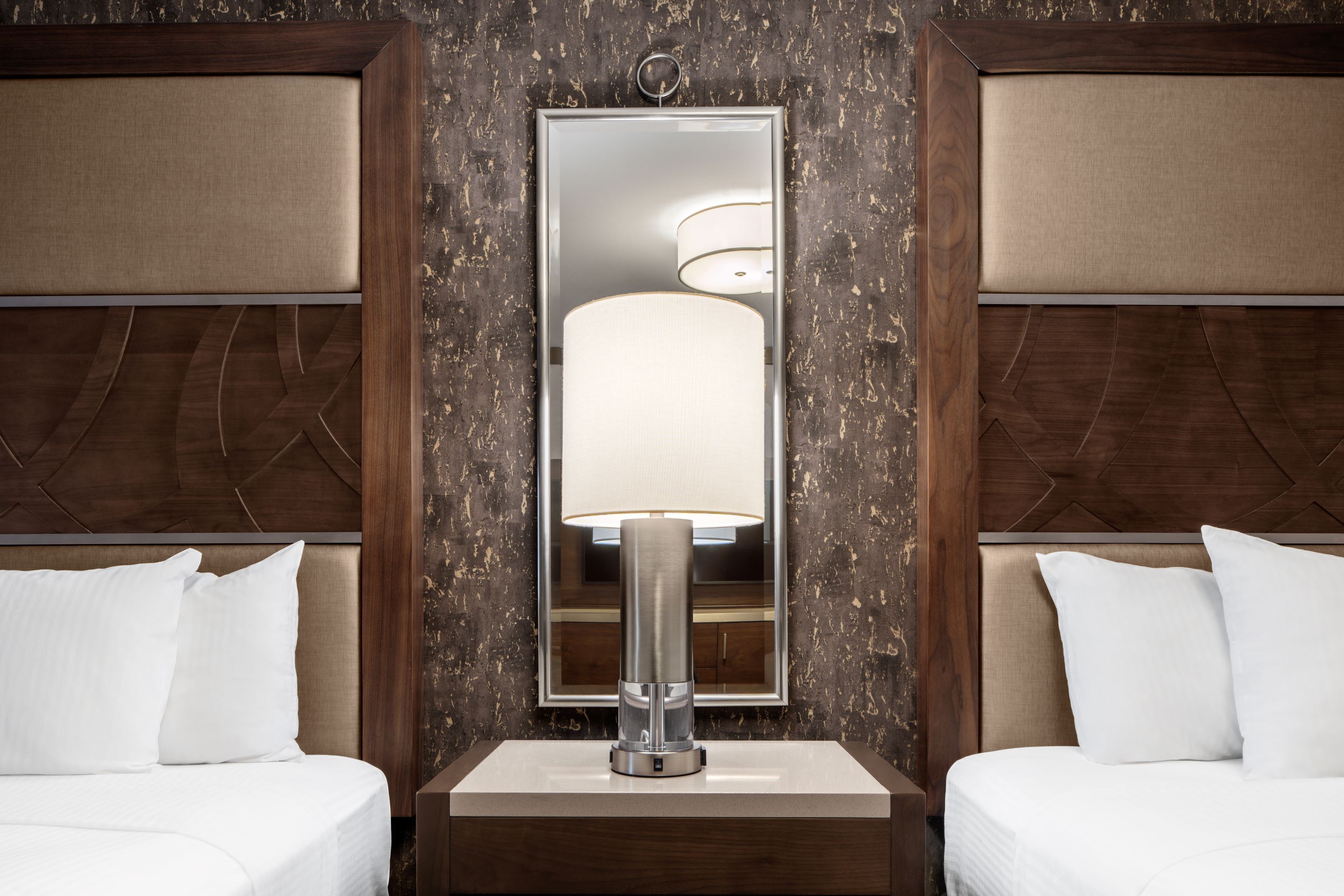 A hotel room with a large mirror, two beds with white linens, a bedside table with a lamp, and a wooden headboard.