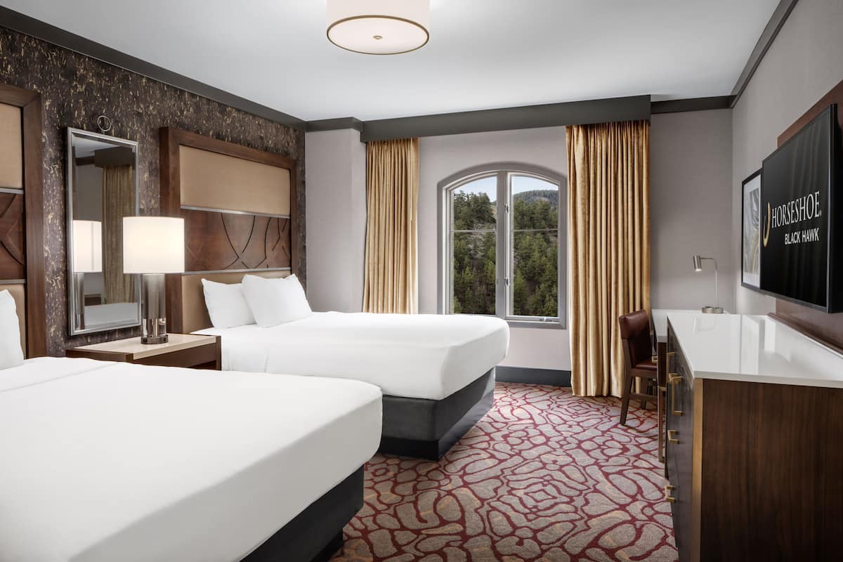 A hotel room with two beds, a large mirror, a TV, a desk, and a window with curtains.