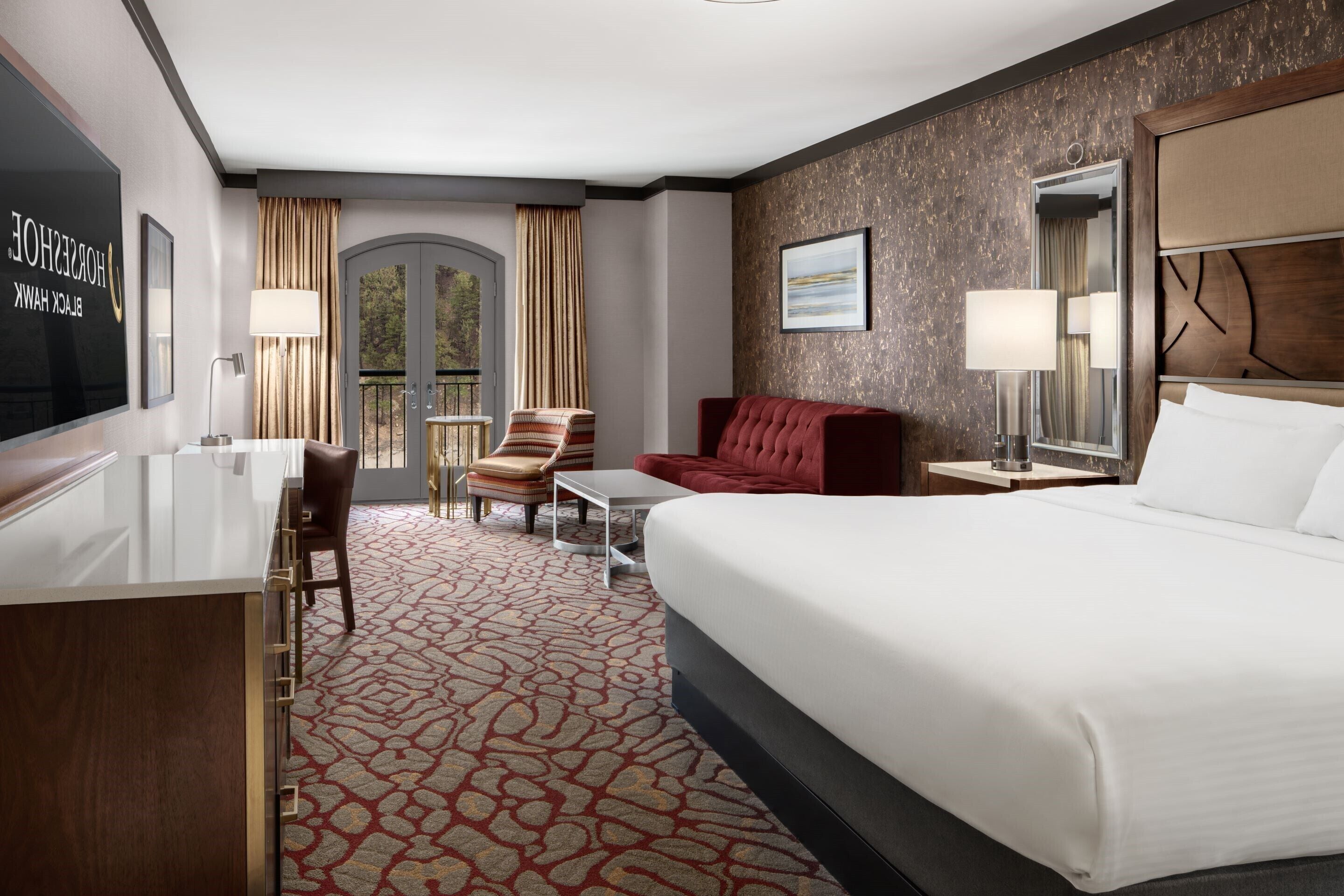 Habitación de hotel con cama, escritorio, silla, sofá rojo, una mesita y ventana con cortinas.