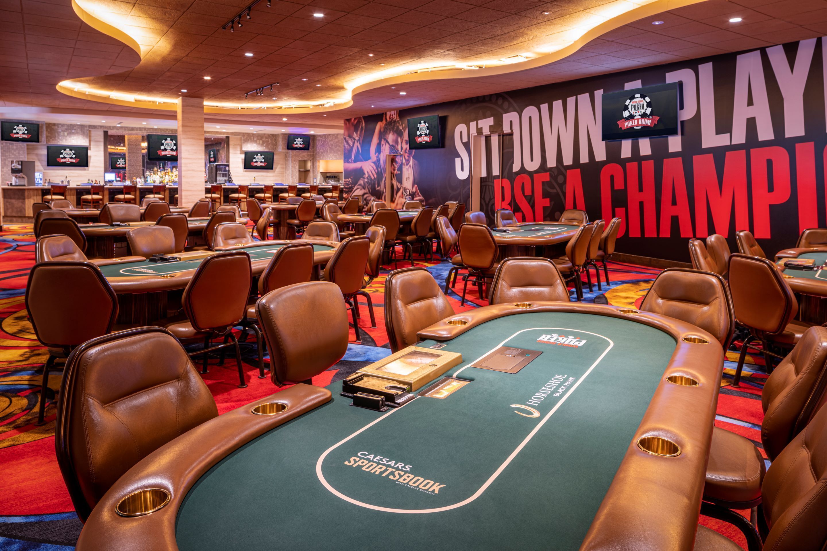 Um casino com várias mesas de blackjack, uma tela grande exibindo 'SITTOWN PLAY TO WIN A CHAMPION' e o logotipo do Caesars Sportsbook nas mesas.