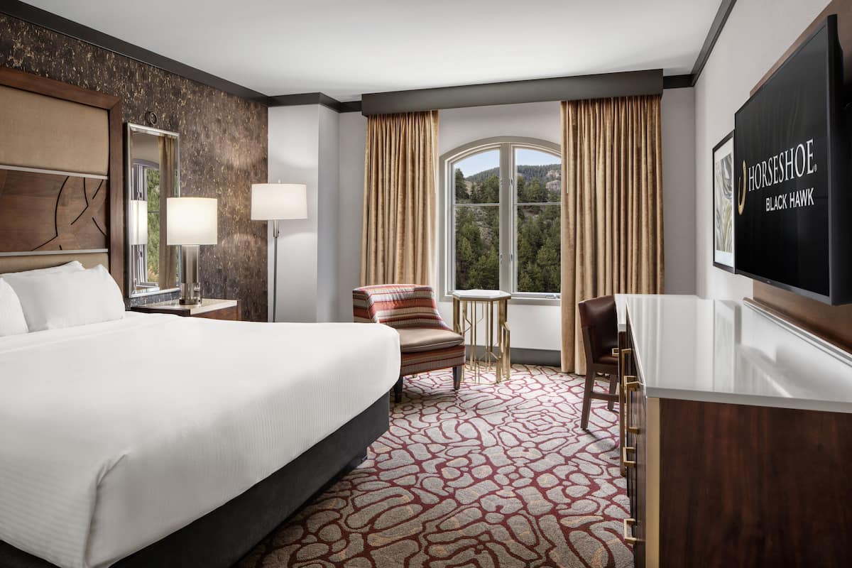 A hotel room with a large bed, a desk, a chair, a small table, a TV, and a window with curtains.
