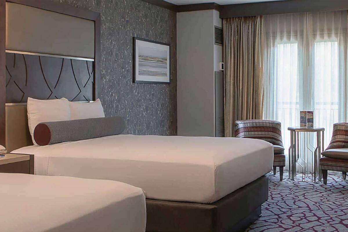 A hotel room with two beds, a bedside table with a lamp, a chair, and a small table.